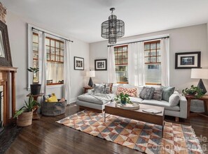 A living room with a sofa, coffee table, and a patterned rug. There are two windows with curtains, a fireplace, and a hanging light fixture. -  Historic Montford house - Walk to downtown Sleeps 6 (Asheville)