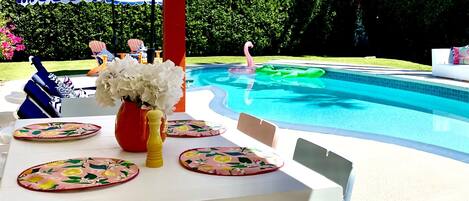Zona de piscina con una mesa dispuesta para comer, rodeada de sombrillas a rayas azules y blancas.