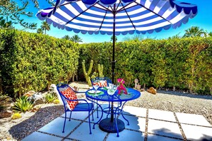 En udendørs terrasse med et rundt bord, blå stole og en stribet parasol.