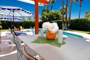 Et poolområde med liggestole, et bord med en blomstervase, og en blå-hvid stribet parasol.