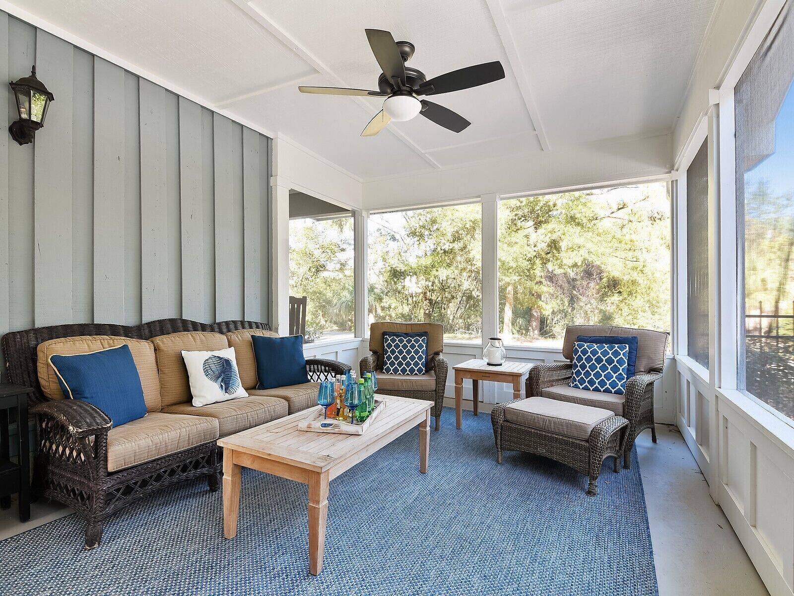 A covered outdoor seating area with a ceiling fan, wicker furniture, and a view of trees through large windows.