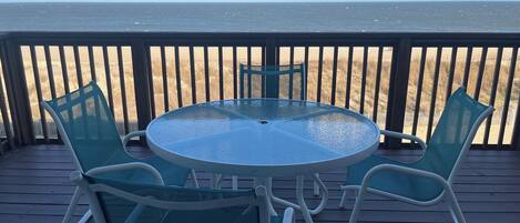 A round glass table with four chairs on a wooden deck, overlooking a beach with a wooden fence.