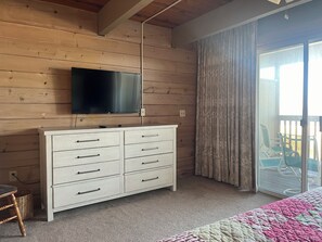 A room with a wooden wall, a white dresser with drawers, a flat-screen TV mounted above it, a bed with a pink and white quilt, and a sliding glass door leading to an outdoor area with chairs. - 4 Bedroom 2 1/2 Bath Ocean Front Townhouse with Outdoor Pool (Ocean City)