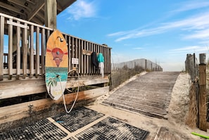 Une promenade en bois mène à une zone de plage où se trouve une planche de surf colorée et un panneau indiquant la présence d’un élément aquatique.