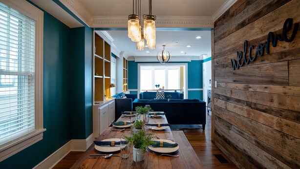 Un comedor con una mesa larga dispuesta para una comida, una pared de madera con un letrero que dice 'welcome' y vista a una sala de estar con sofá y ventana.