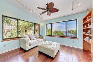 A spacious living room with large windows, a ceiling fan, a white sofa, an ottoman, and a wooden bookshelf.