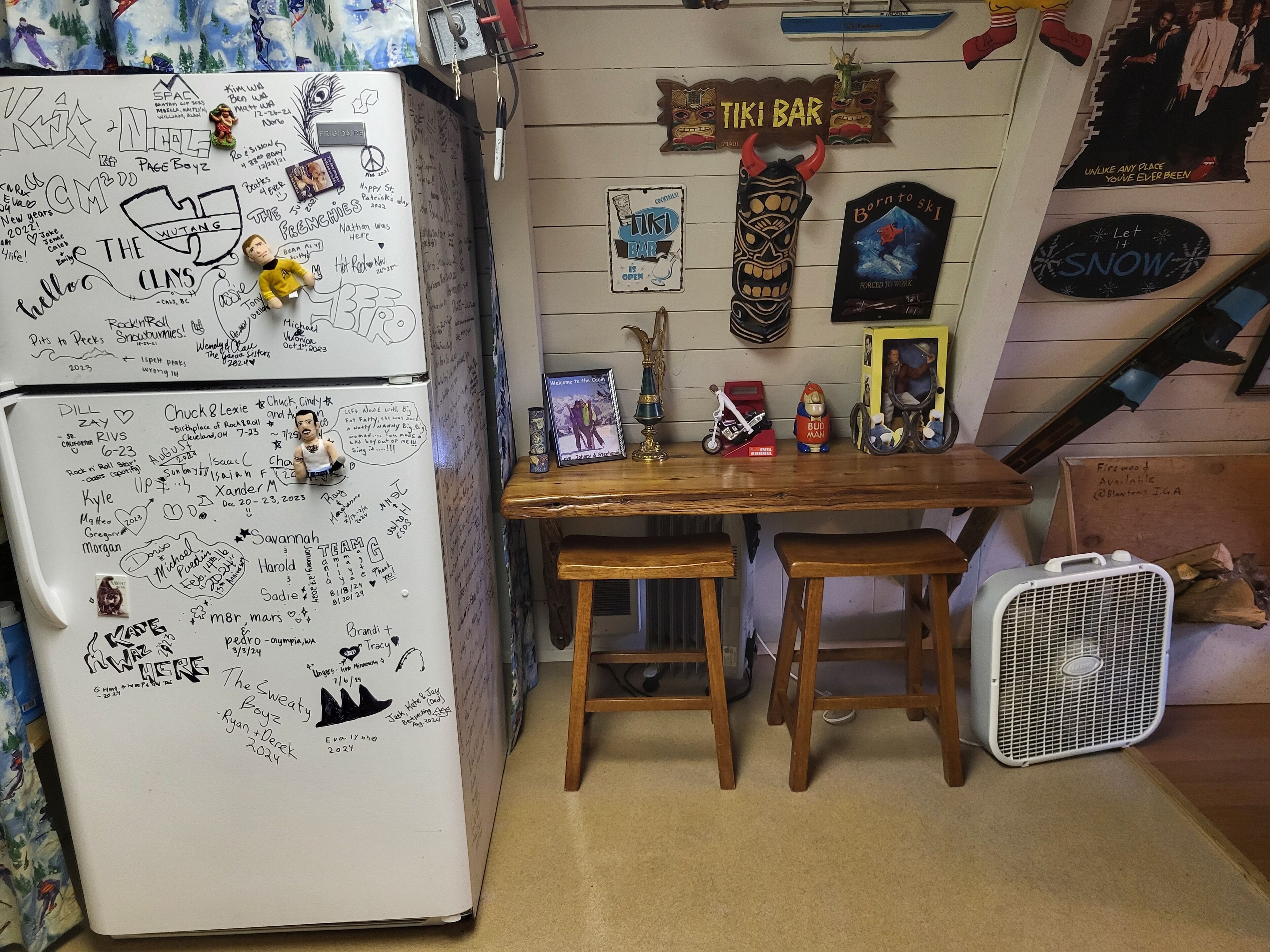 A small kitchen with a refrigerator covered in magnets and notes, a wooden table with chairs, and a fan.
