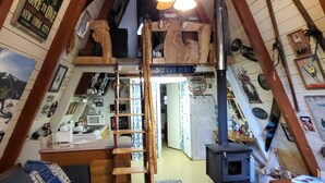 Una habitación estilo loft con techo inclinado, vigas de madera, ventilador de techo, una escalera, kitchenette y un amplio ventanal con vistas al exterior.