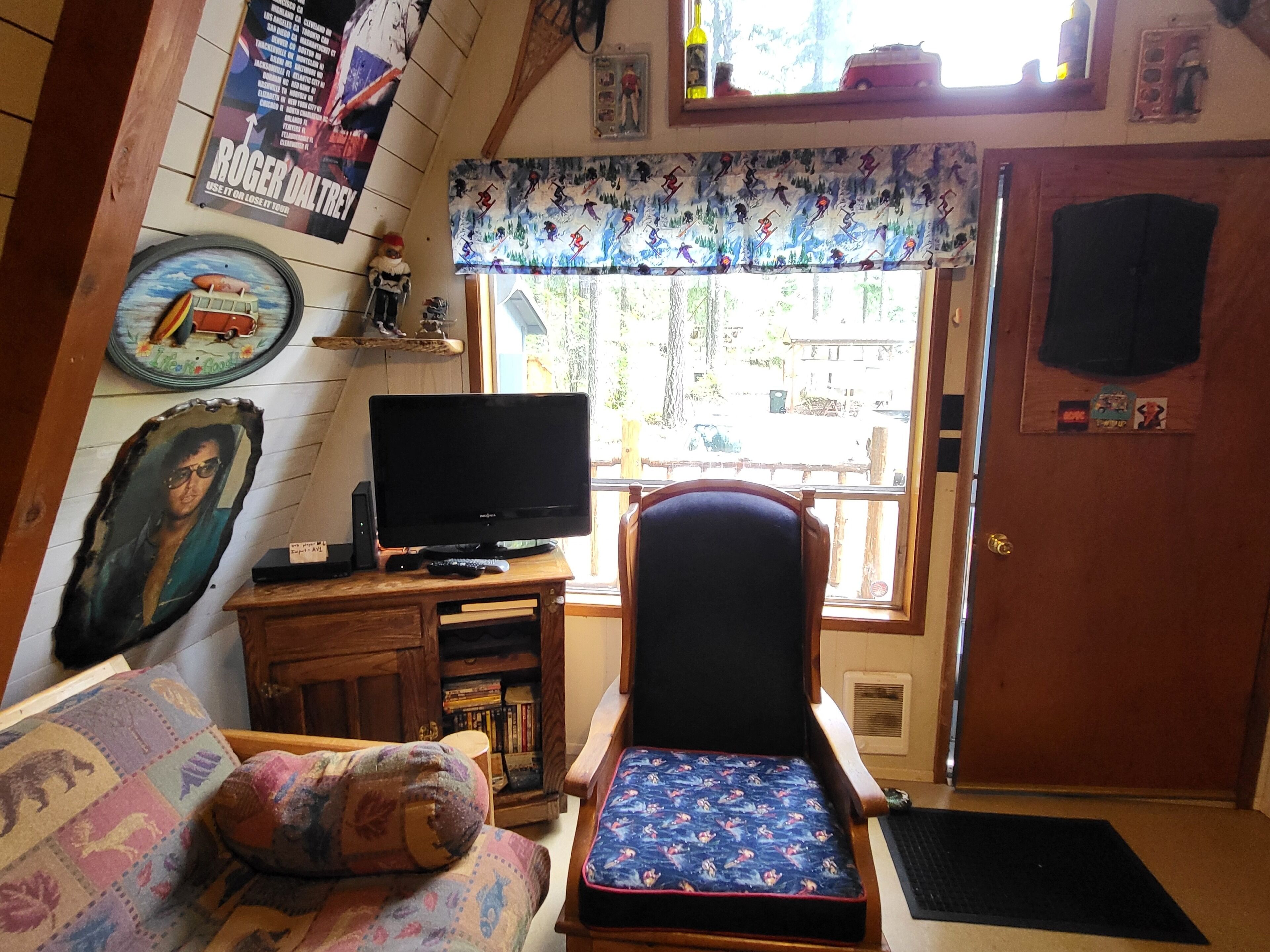 Una sala de estar acogedora con un televisor sobre un mueble de madera, un sillón estampado, una pequeña estantería y un ventanal con un valance decorativo.