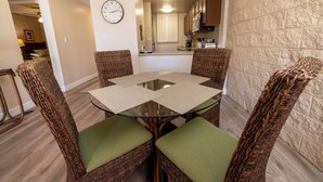 A dining area with a glass-top table, wicker chairs with green cushions, a clock on the wall, and a kitchen in the background. - Oceanview Kanai A Nalu 316 Great Views Split AC Pool Great Value (Wailuku)