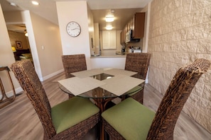 A dining area with a glass-top table, wicker chairs with green cushions, a clock on the wall, and a kitchen in the background. - Oceanview Kanai A Nalu 316 Great Views Split AC Pool Great Value (Wailuku)