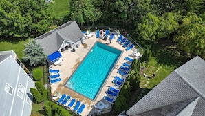 An aerial view of a rectangular swimming pool surrounded by blue lounge chairs, with a covered patio area and a small deck.