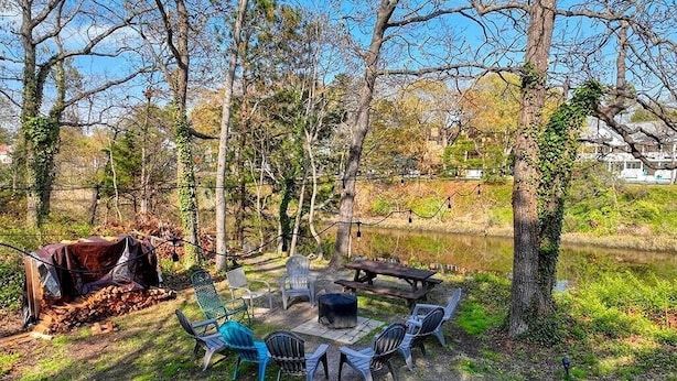 An outdoor seating area with a fire pit, chairs, and a picnic table surrounded by trees and a body of water.