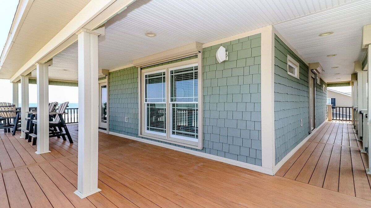 A covered outdoor area with a wooden deck, a covered seating area with chairs, and a window with a view.