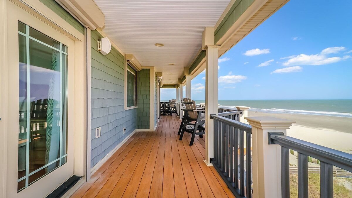 Una terraza de madera con barandilla, vista a la playa y mobiliario exterior.