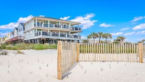 Een huis aan het strand met een twee verdiepingen tellend balkon, een houten omheining en palmbomen.