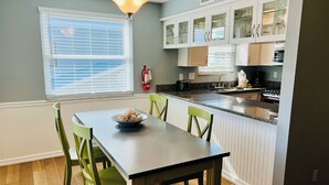A kitchen with a dining table, chairs, and a bowl of fruit. There is a window with blinds, a fire extinguisher, and cabinets with glass doors. - Live like a local. Heated Pool, steps to the Beach. 2nd floor unit. Unit B (Indian Rocks Beach)