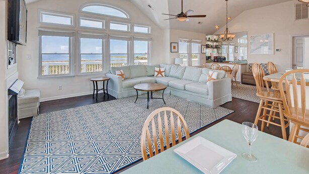 A spacious living room with a large window overlooking the sea, a cozy seating area, and a dining space with wooden chairs and a table. - Semi-Soundfront Dog Friendly w/ Pool, Hot Tub, Game Room, Community Boat Ramp (Salvo)