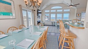 A dining area with a long table set for six, wooden chairs, a patterned rug, and a view of the sea through large windows. - Semi-Soundfront Dog Friendly w/ Pool, Hot Tub, Game Room, Community Boat Ramp (Salvo)