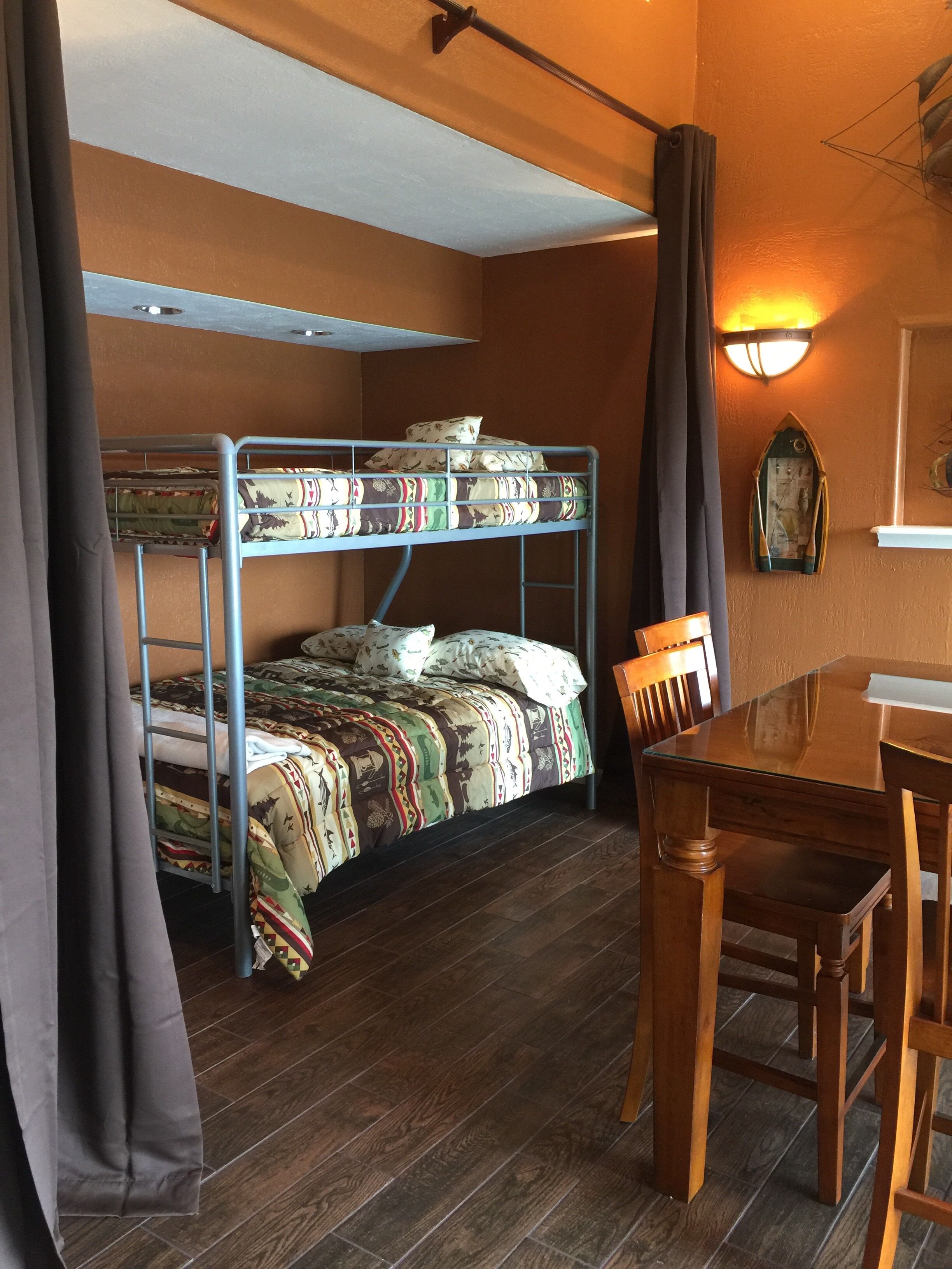 A bunk bed with patterned bedding, a wooden table with chairs, and a wall-mounted light fixture.