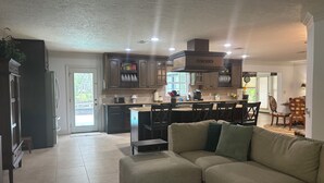 Una cocina moderna con armarios de madera oscura, una isla central y un horno empotrado. En la zona de estar, hay un sofá seccional color beige.
