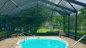 Una piscina interior con cerramiento de vidrio, suelo de piedra y vista a un jardín ajardinado.