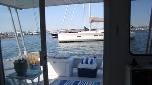 Cubierta de barco con zona de estar acolchada, una pequeña mesa con un cuenco de fruta y vistas a otros barcos en el puerto deportivo.