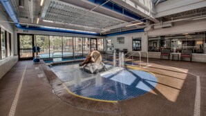 Une piscine intérieure moderne dotée d’un élément aquatique central, de grandes fenêtres et d’un plafond haut laissant apparaître les conduits de ventilation.