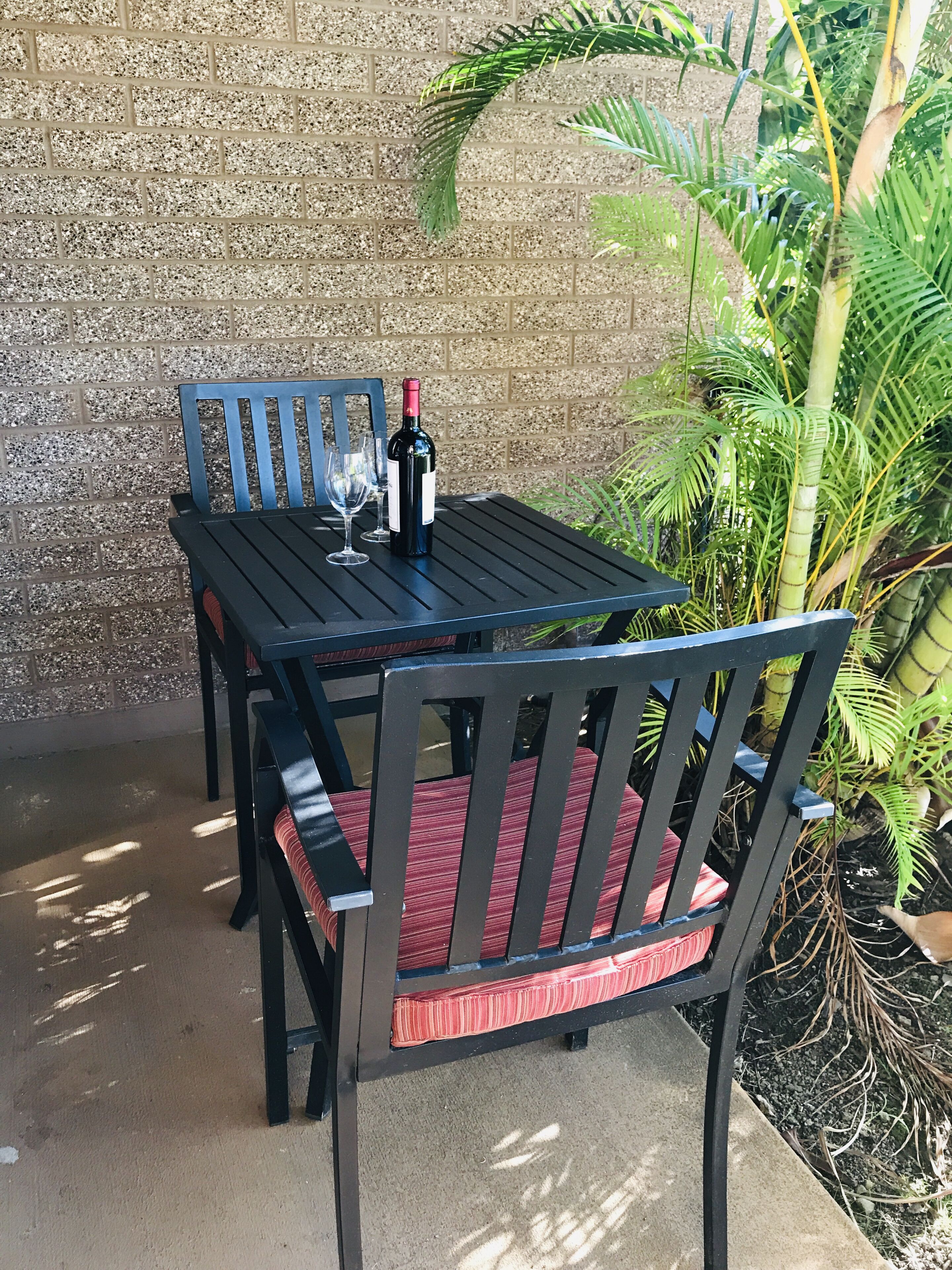 Une table et des chaises de patio, accompagnées d’une bouteille de vin et de verres, sont disposées devant un mur de briques, avec de la verdure en arrière-plan.