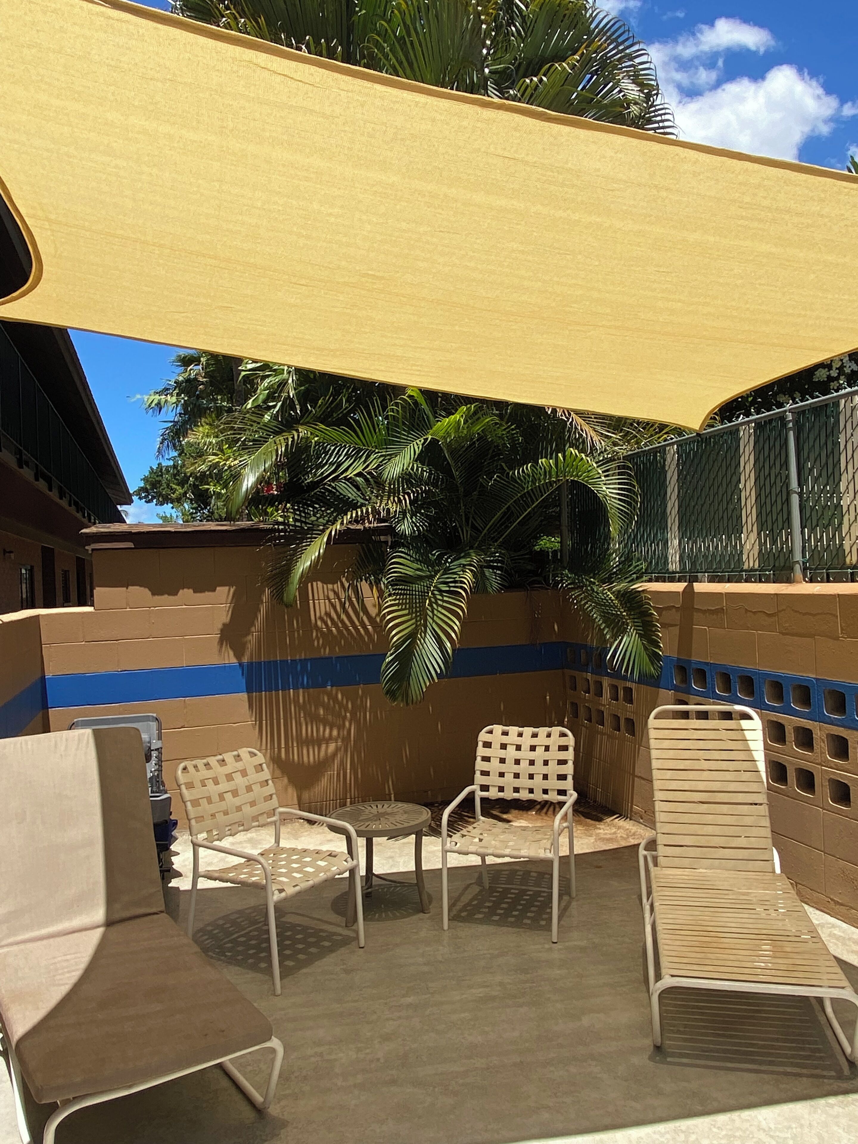 Un área de estar al aire libre con cuatro sillones y una mesa, protegida por un toldo amarillo, con palmeras y una pared de ladrillo al fondo.