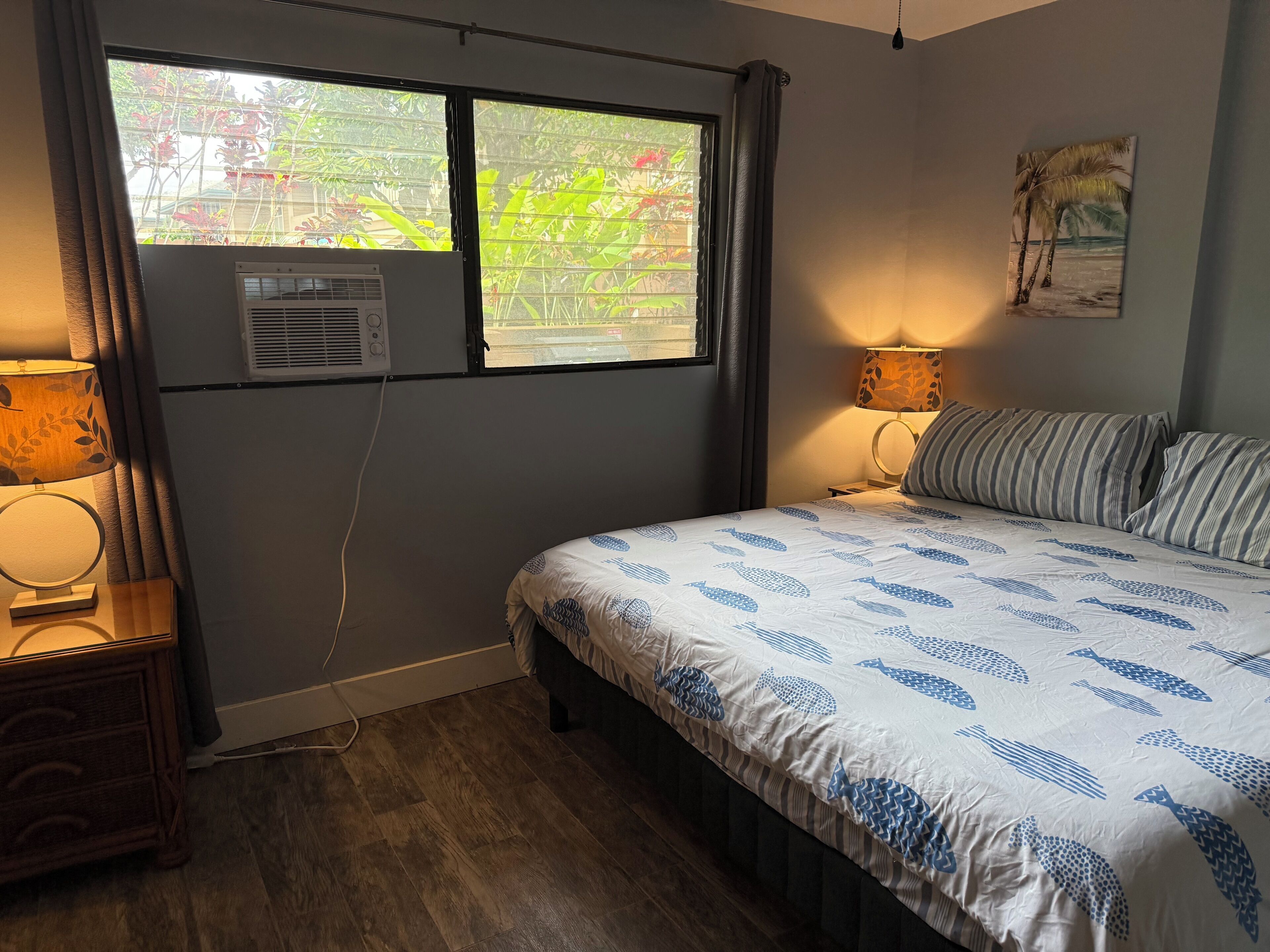 Un dormitorio con cama, dos lámparas de noche, ventana con aire acondicionado y un cuadro colgado en la pared.