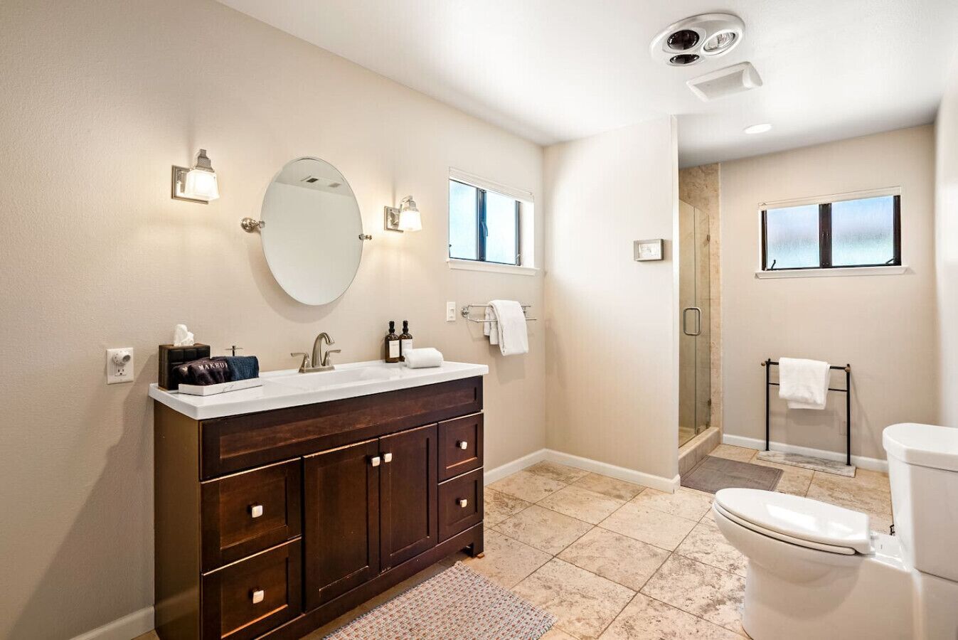 Un baño moderno con un espejo grande, un tocador con muebles de madera oscura, un lavamanos blanco y un inodoro. Hay dos ventanas que aportan luz natural y una zona de ducha con puerta de cristal.