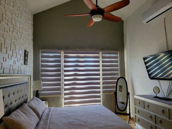 A bedroom with a bed, a ceiling fan, a TV, a dresser, and a window with blinds. - CASA BORINQUEN VACATION RENTAL (Aguadilla)