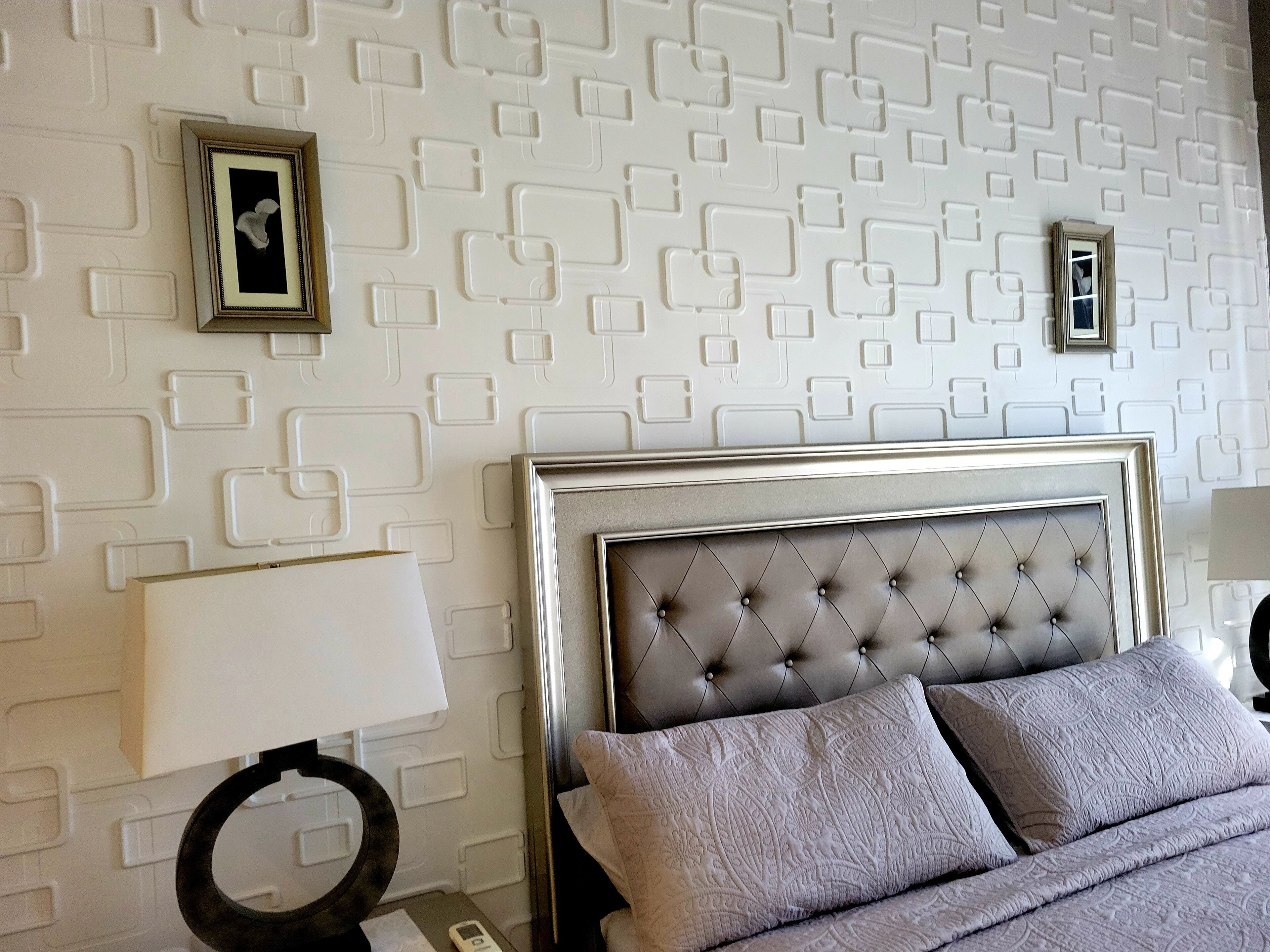 A neatly arranged bedroom with a tufted headboard, a bedside table with a lamp, and patterned wall decor.
