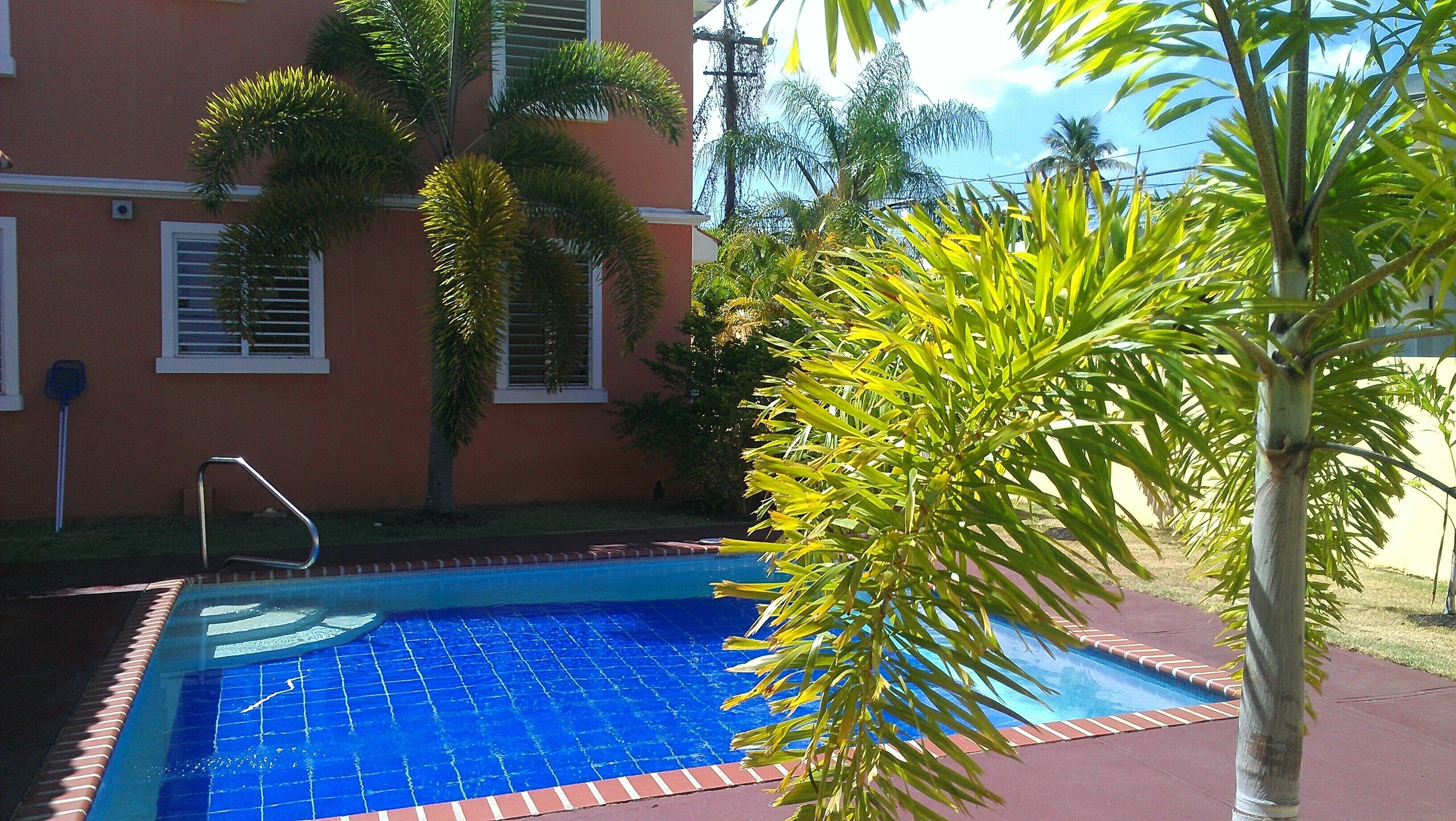 Una piscina con solárium de baldosas, rodeada de palmeras y un edificio con ventanas.