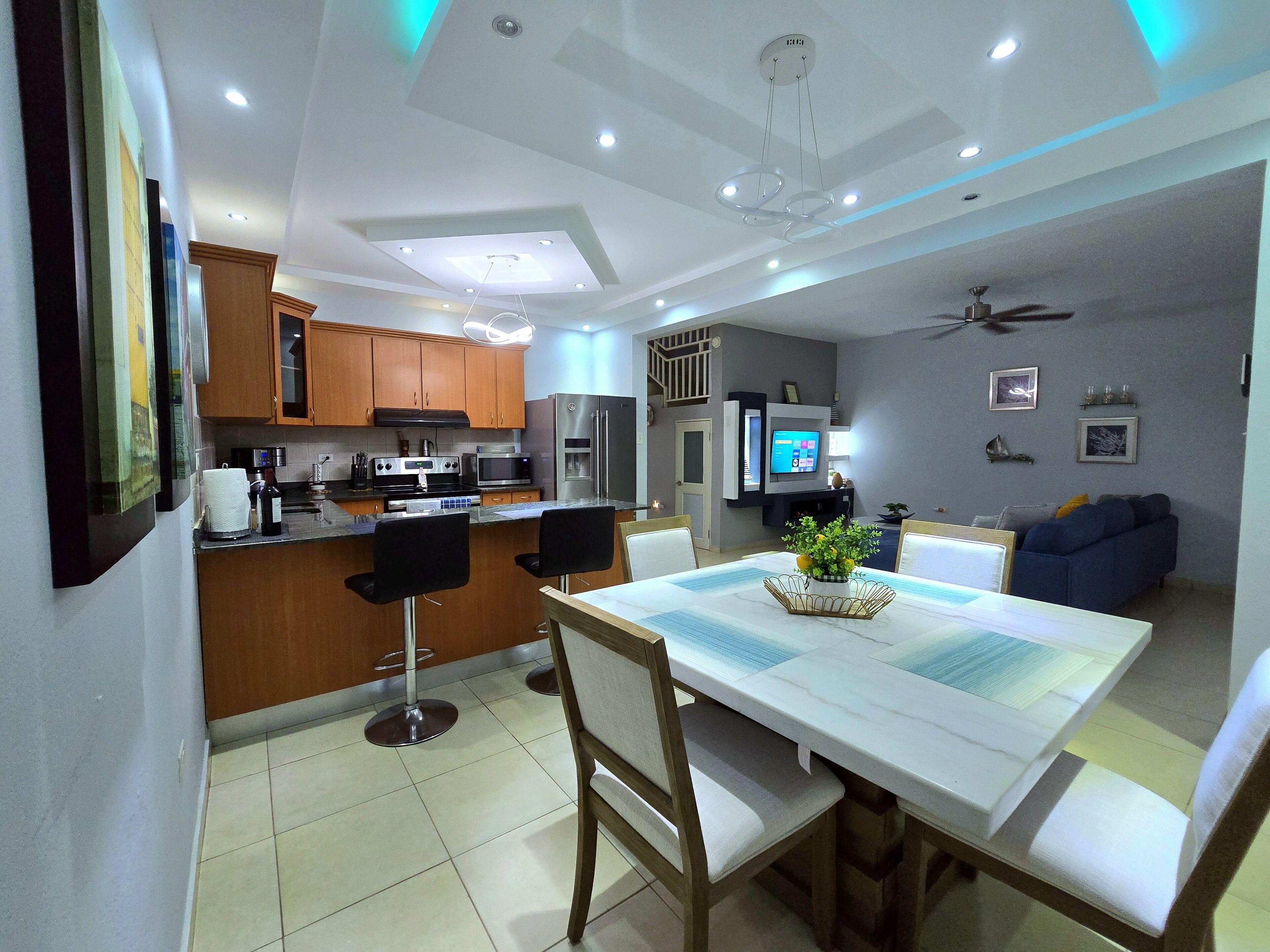 A modern kitchen with wooden cabinets, a dining area with a white table, and a living room with a blue sofa.
