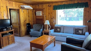 A cozy room with wooden walls, a television on a wooden stand, a grey sofa with animal-themed cushions, a blue armchair, and a wooden coffee table. - Rustic Charm Meets Modern Comfort, Steps from Yellowstone's West Entrance (West Yellowstone)