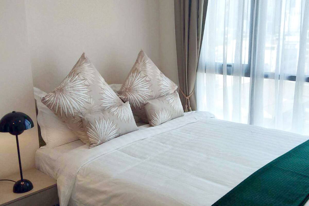 A neatly made bed with white bedding and a green throw, a bedside lamp, and a window with sheer curtains.