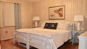 A bedroom with a wooden paneled wall, a bed with a patterned bedspread, two bedside tables with lamps, and a painting above the bed. - Perfect for multi-family gatherings! (Oak Island)