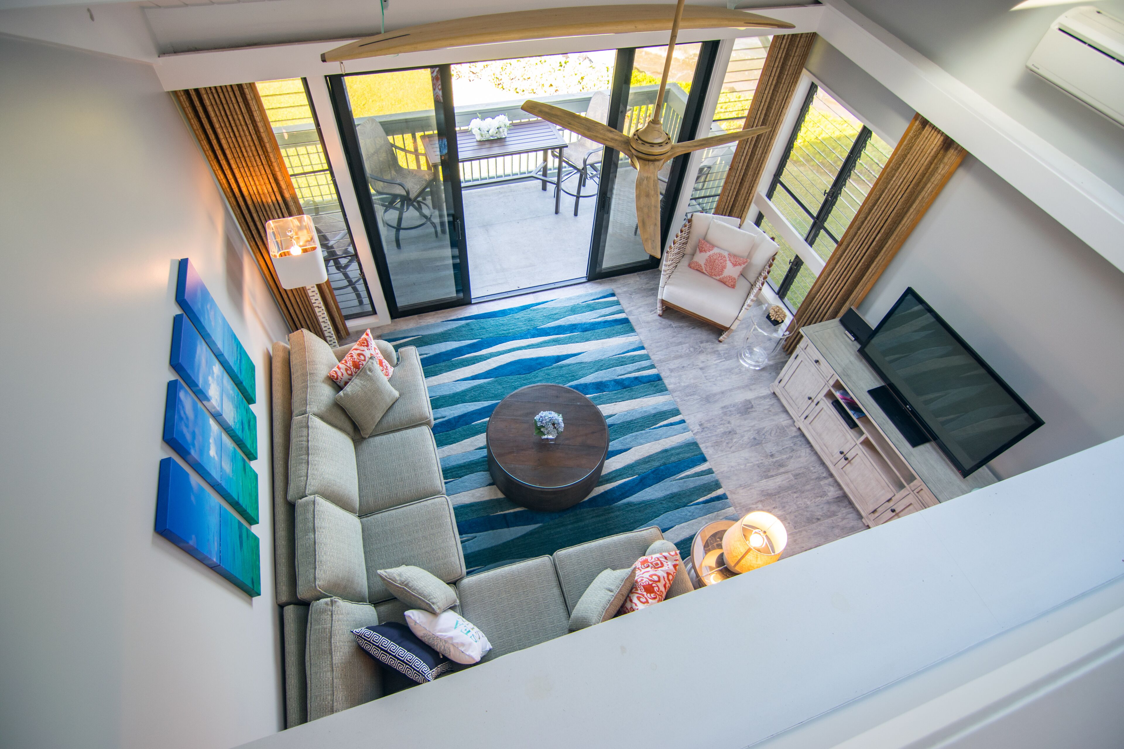 A modern living room with a sectional sofa, a coffee table, and a TV. There is a large window with curtains, a ceiling fan, and a view of the outside patio.