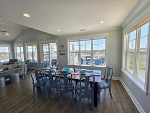 Un espace repas avec une grande table, des chaises bleues et une vue sur l’extérieur grâce à de grandes fenêtres.