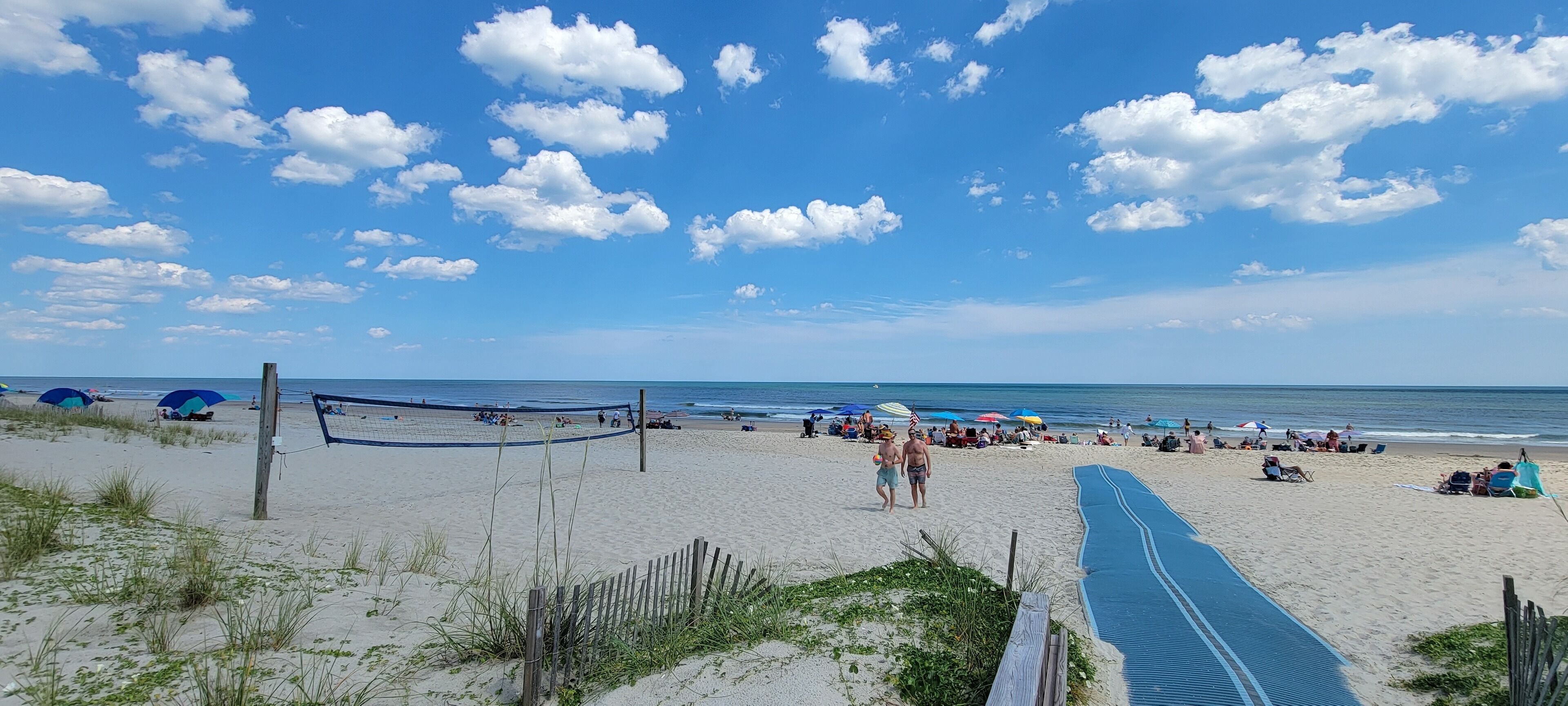 A beach with a volleyball net, umbrellas, and people.