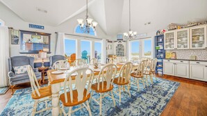 A dining area with a large table, multiple chairs, and a view of the beach through large windows.