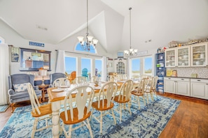 A dining area with a large table, multiple chairs, and a view of the beach through large windows.
