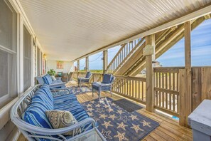 Uma área de estar coberta ao ar livre com vista para a praia, com um sofá, cadeiras e um deck de madeira.