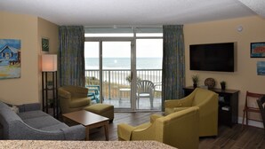 Una sala de estar con un sofá, sillones, una mesa de centro y un televisor. Hay una puerta corrediza de vidrio que da a un balcón con vistas a la playa.