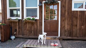 Una casa de madera con un perro de pie sobre una plataforma de madera frente a la puerta.