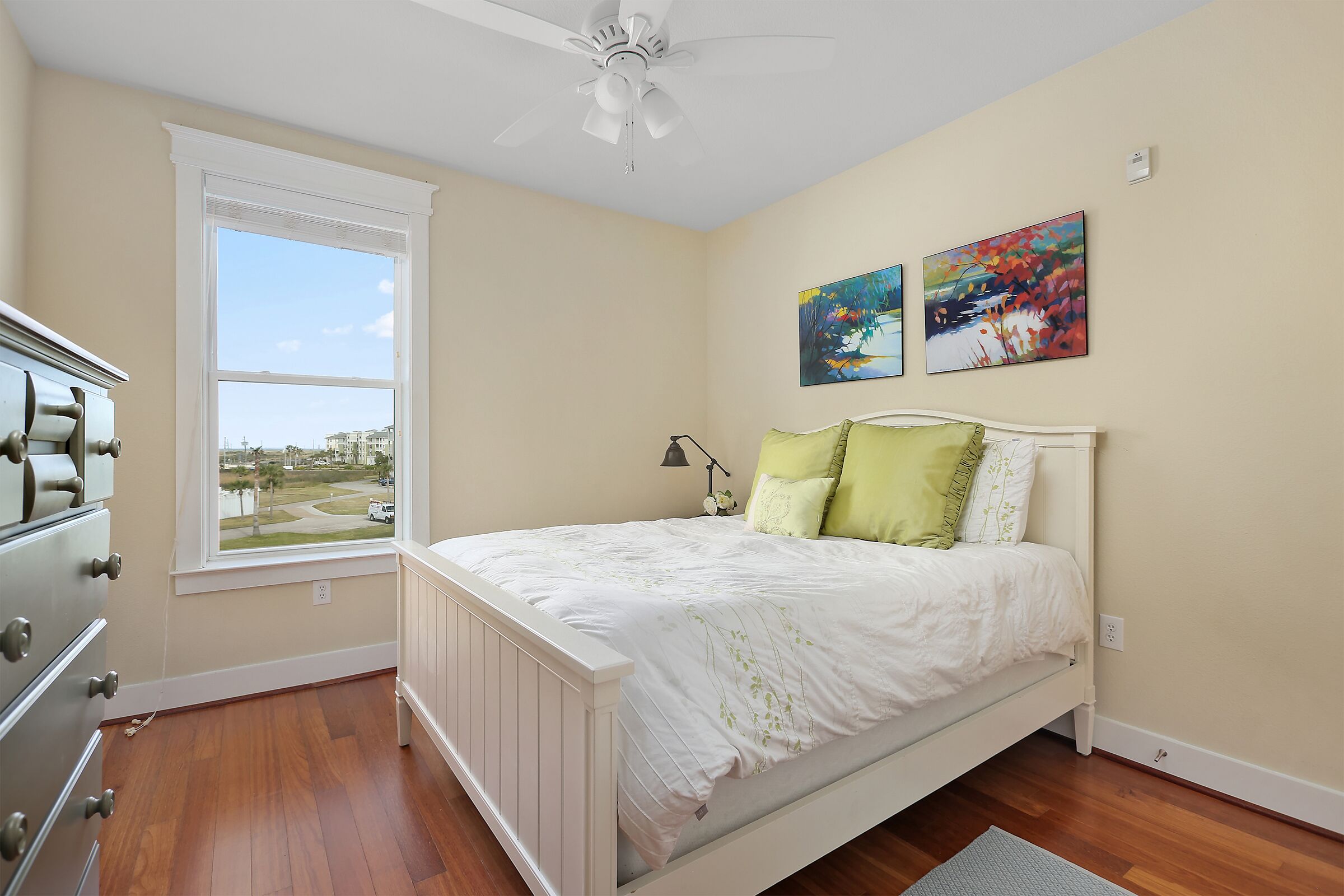 A bedroom with a bed, a window, a ceiling fan, and artwork on the wall.