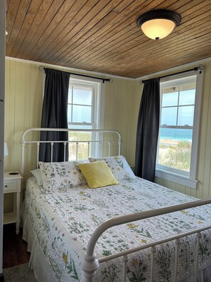 Une chambre avec un plafond en bois, un lit recouvert d’une literie à motifs floraux, deux fenêtres avec des rideaux et une vue sur la plage.