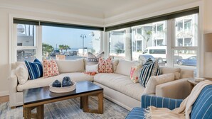 A modern living room with a sectional sofa, a coffee table, and large windows offering a view of the street and buildings. - LOCATION!!!  7 Bedrooms - VIEW - Steps to Sand+Ocean+Pier+Downtown PISMO BEACH! (Pismo Beach)